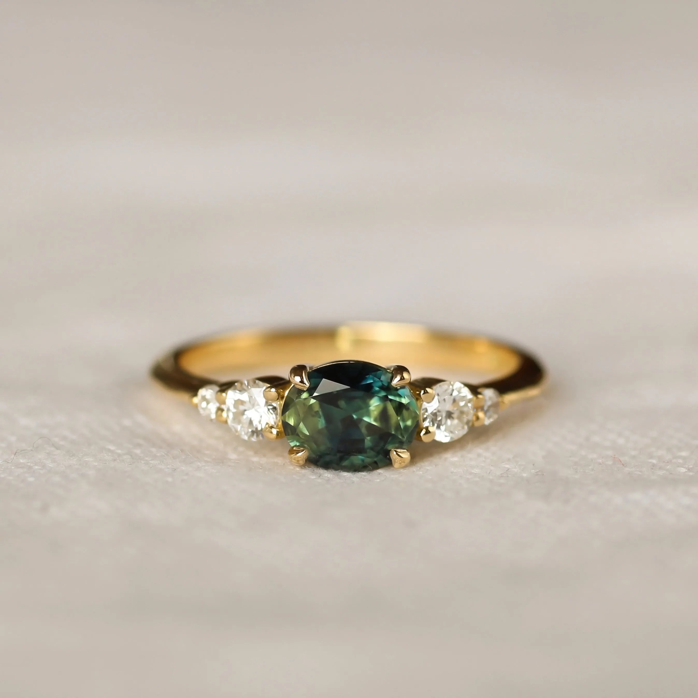 bi colour sapphire engagement ring with east-west oval sapphire with blue and green colours, round diamonds on a neutral surface