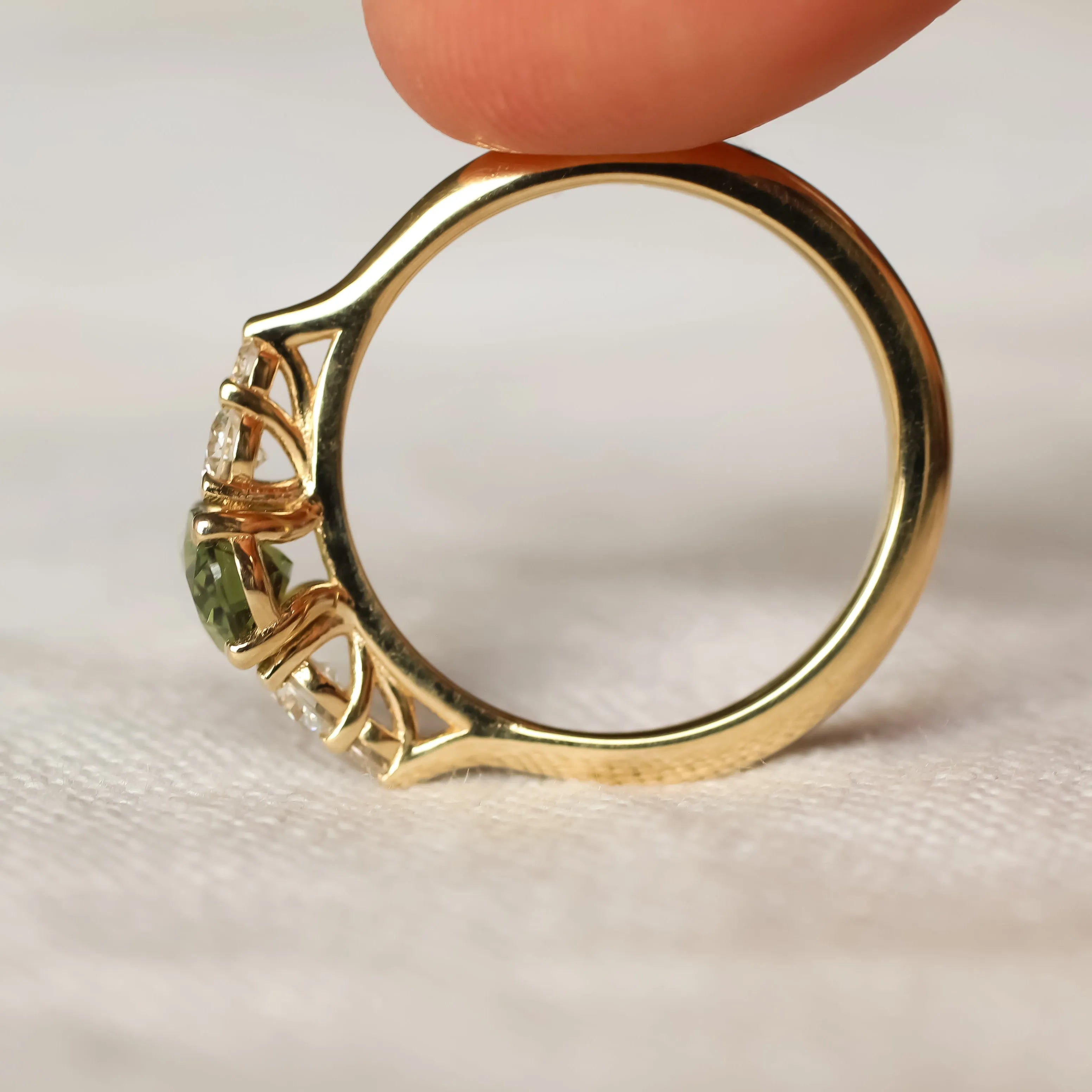 Gold wedfit engagement ring with a green gemstone held between fingers on a neutral background