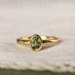 gold engagement ring with textured organic band and green oval sapphire surrounded by gold on a neutral fabric