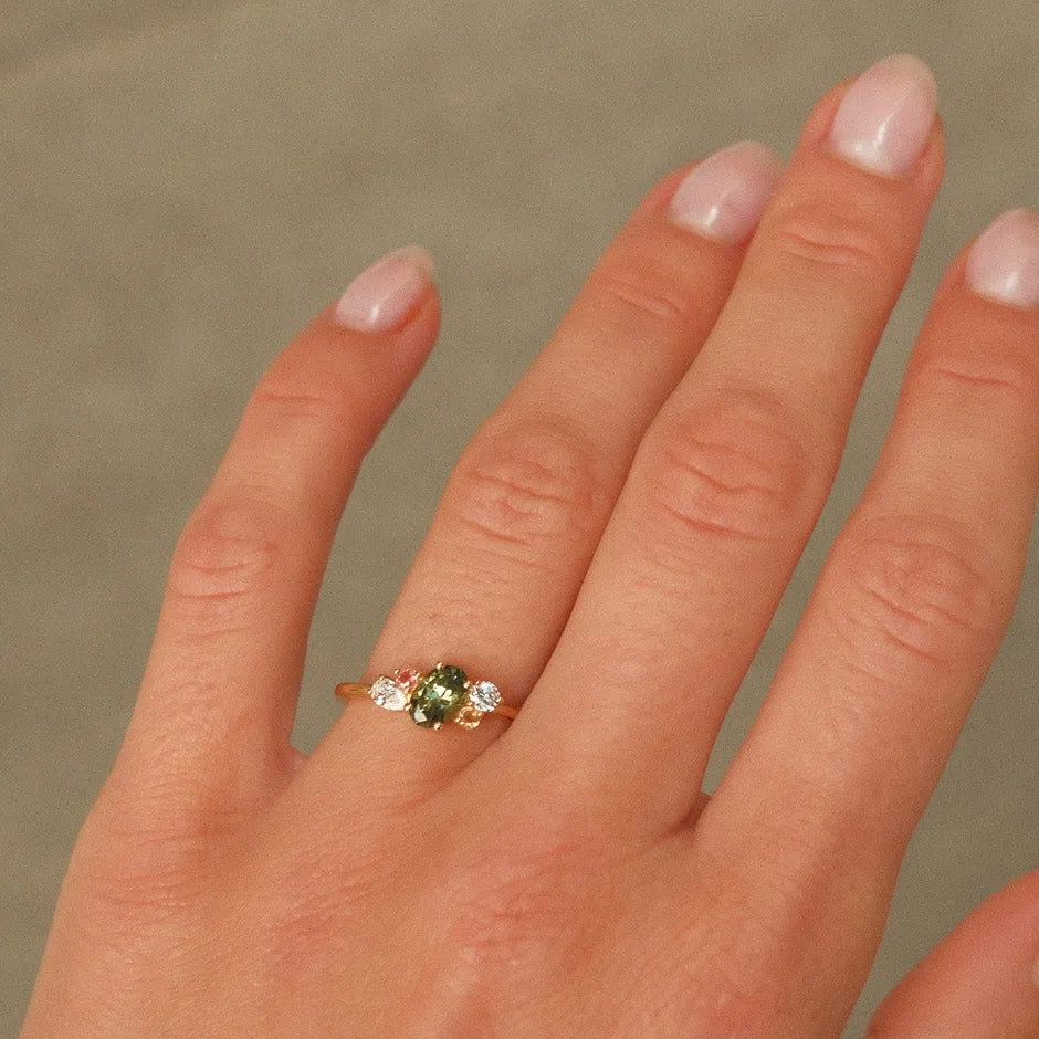 Hand wearing a ring with a colourful engagement ring with green, pink and yellow gemstones on a neutral background