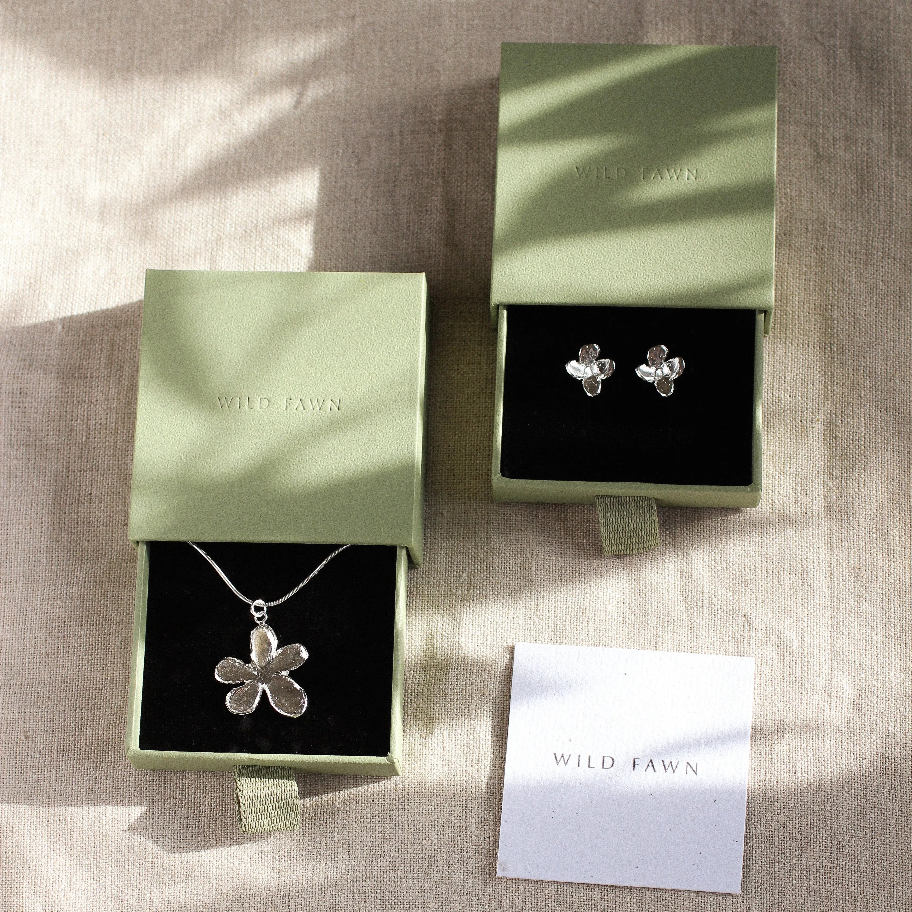 Silver flower necklace and earrings in open green boxes on a textured surface with 'Wild Pawn' branding.