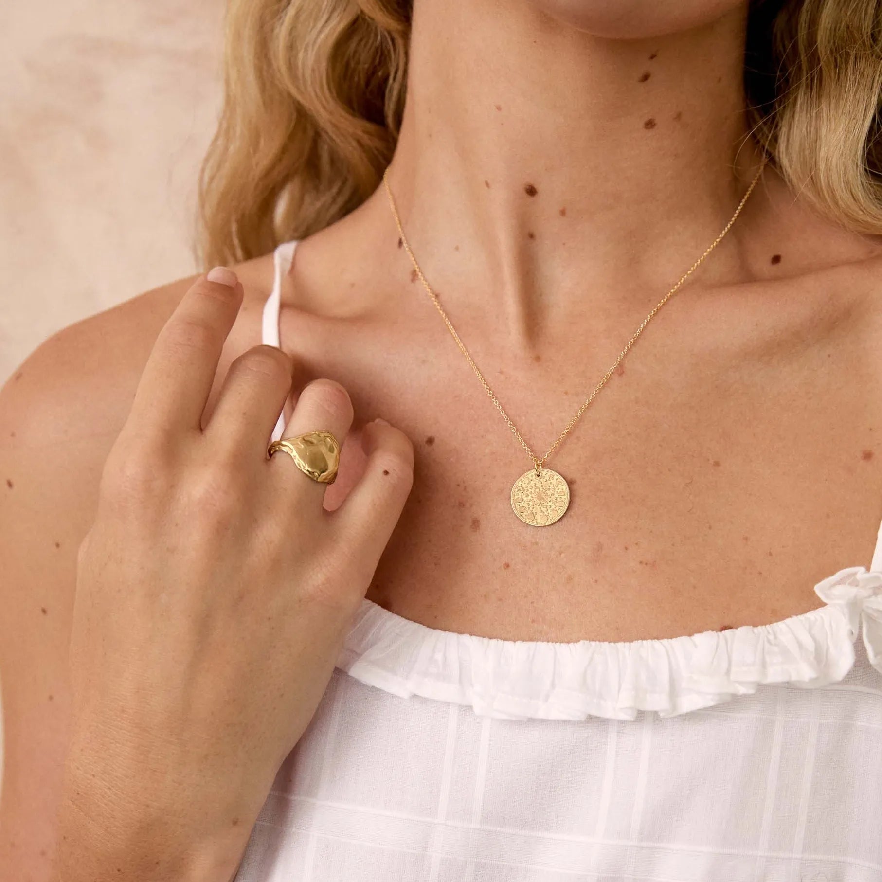 A close-up lifestyle shot of a model wearing a delicate gold coin pendant necklace and a gold ring, styled with a white ruffled spaghetti strap top against a soft beige background.