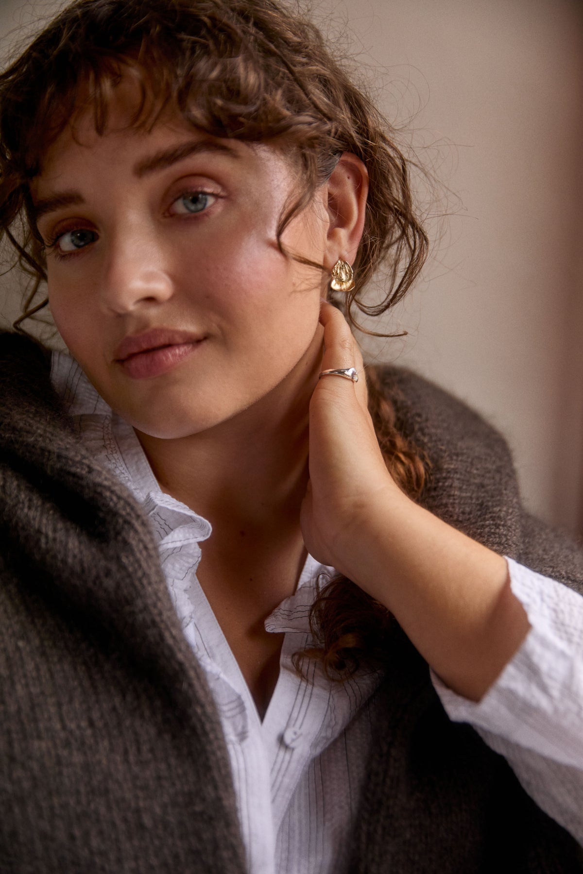 A close-up portrait of a woman with curly brown hair wearing a gold hoop earring and a silver ring, wrapped in a dark gray knit sweater over a white collared shirt.