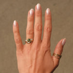Hand wearing a ring with a green gemstone with a chunky curved wedding ring against a neutral background