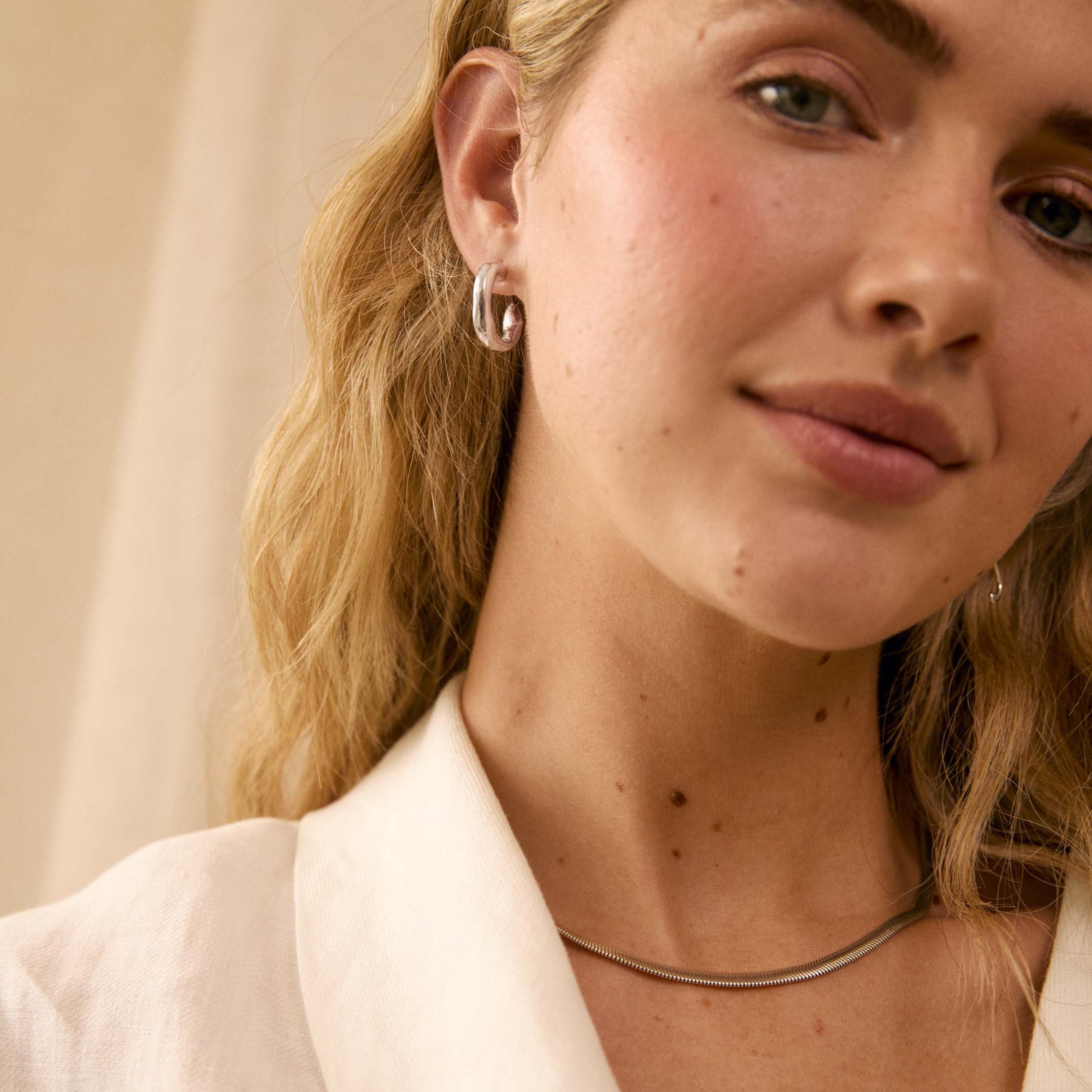 A close-up lifestyle shot of a woman with blonde hair wearing a silver snake chain necklace and small silver hoop earrings, dressed in a white blazer against a soft beige background.