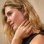 A close-up portrait of a woman with blonde hair wearing gold rings and a gold hoop earring, posing with her hand near her neck against a soft beige background.