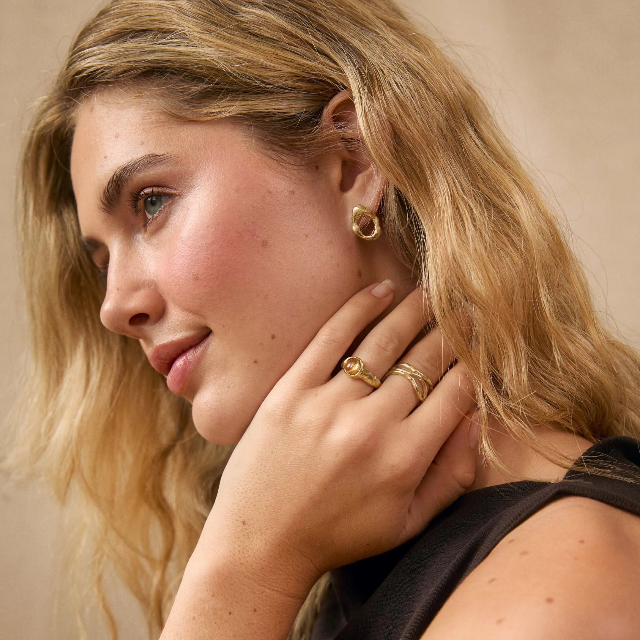 A close-up portrait of a woman with blonde hair wearing gold rings and a gold hoop earring, posing with her hand near her neck against a soft beige background.