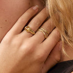A close-up portrait of a woman with blonde hair wearing gold rings and a gold hoop earring, posing with her hand near her neck against a soft beige background.