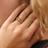 A close-up portrait of a woman with blonde hair wearing gold rings and a gold hoop earring, posing with her hand near her neck against a soft beige background.