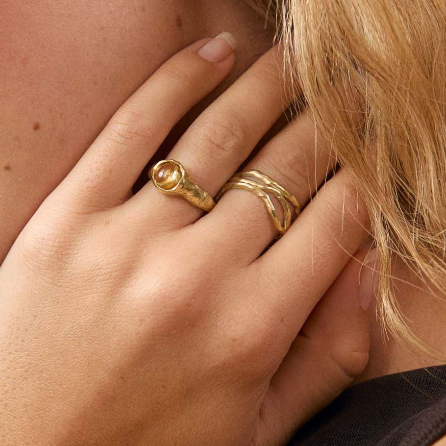A close-up portrait of a woman with blonde hair wearing gold rings and a gold hoop earring, posing with her hand near her neck against a soft beige background.