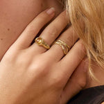 A close-up portrait of a woman with blonde hair wearing gold rings and a gold hoop earring, posing with her hand near her neck against a soft beige background.