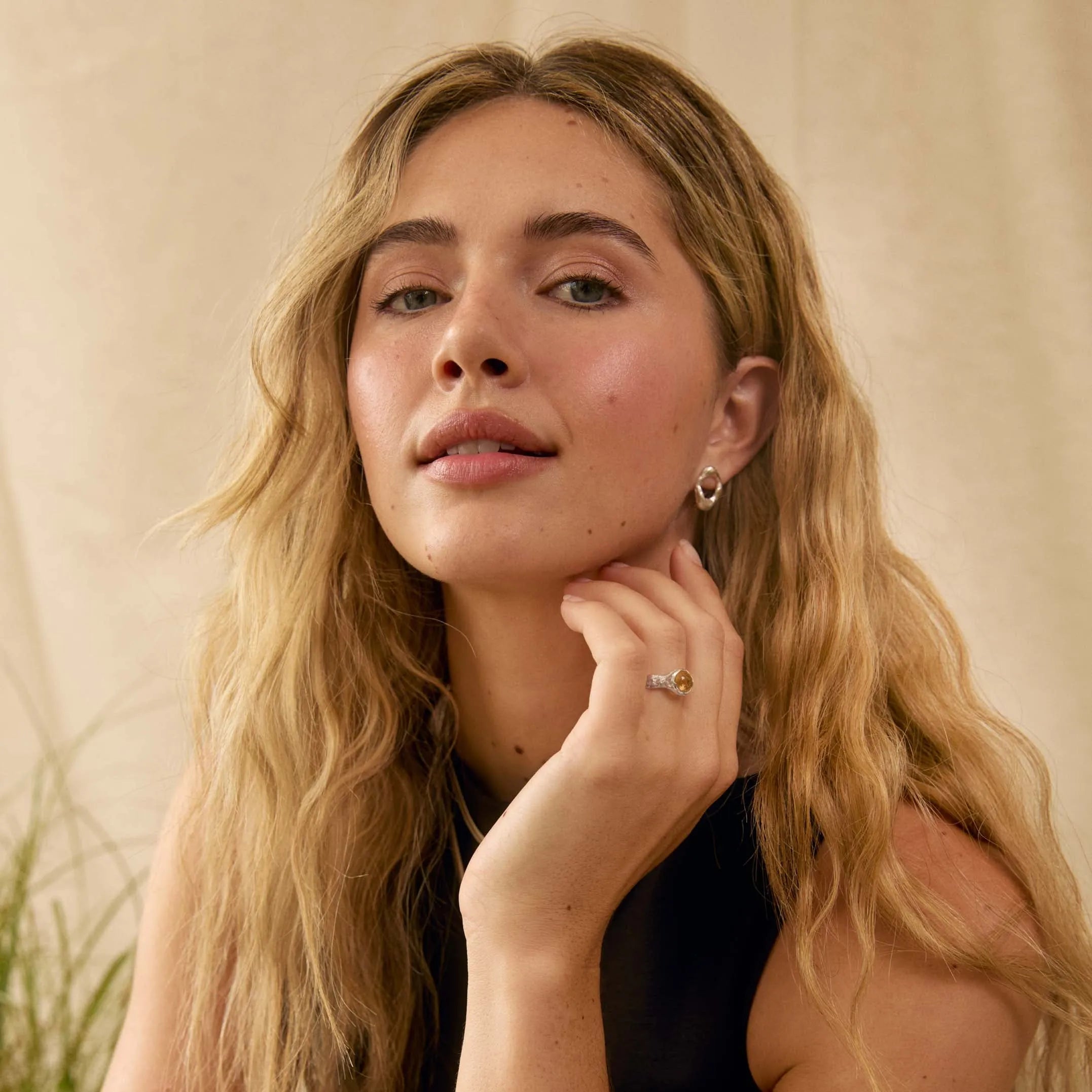 A woman with long wavy blonde hair wearing a black sleeveless top, resting her chin on her hand. She is wearing a silver ring on one finger and a small silver hoop earring, set against a warm beige background with soft lighting.