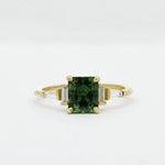 Gold engagement ring with a green cushion cut sapphire and tapered baguetter diamonds on a white background