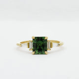 Gold engagement ring with a green cushion cut sapphire and tapered baguetter diamonds on a white background