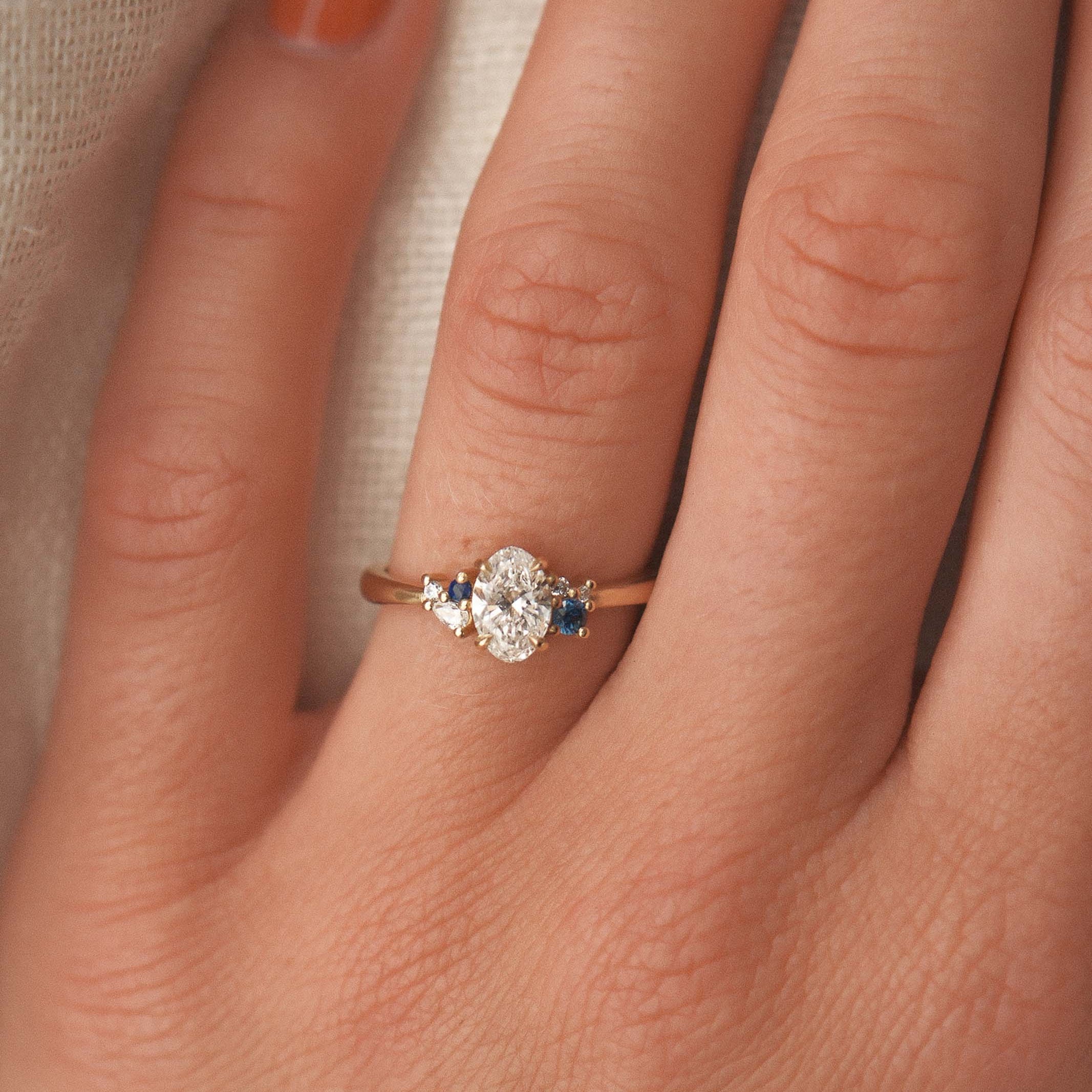 Close-up of a hand wearing an elegant ring with a diamond and blue gemstone on a neutral background.