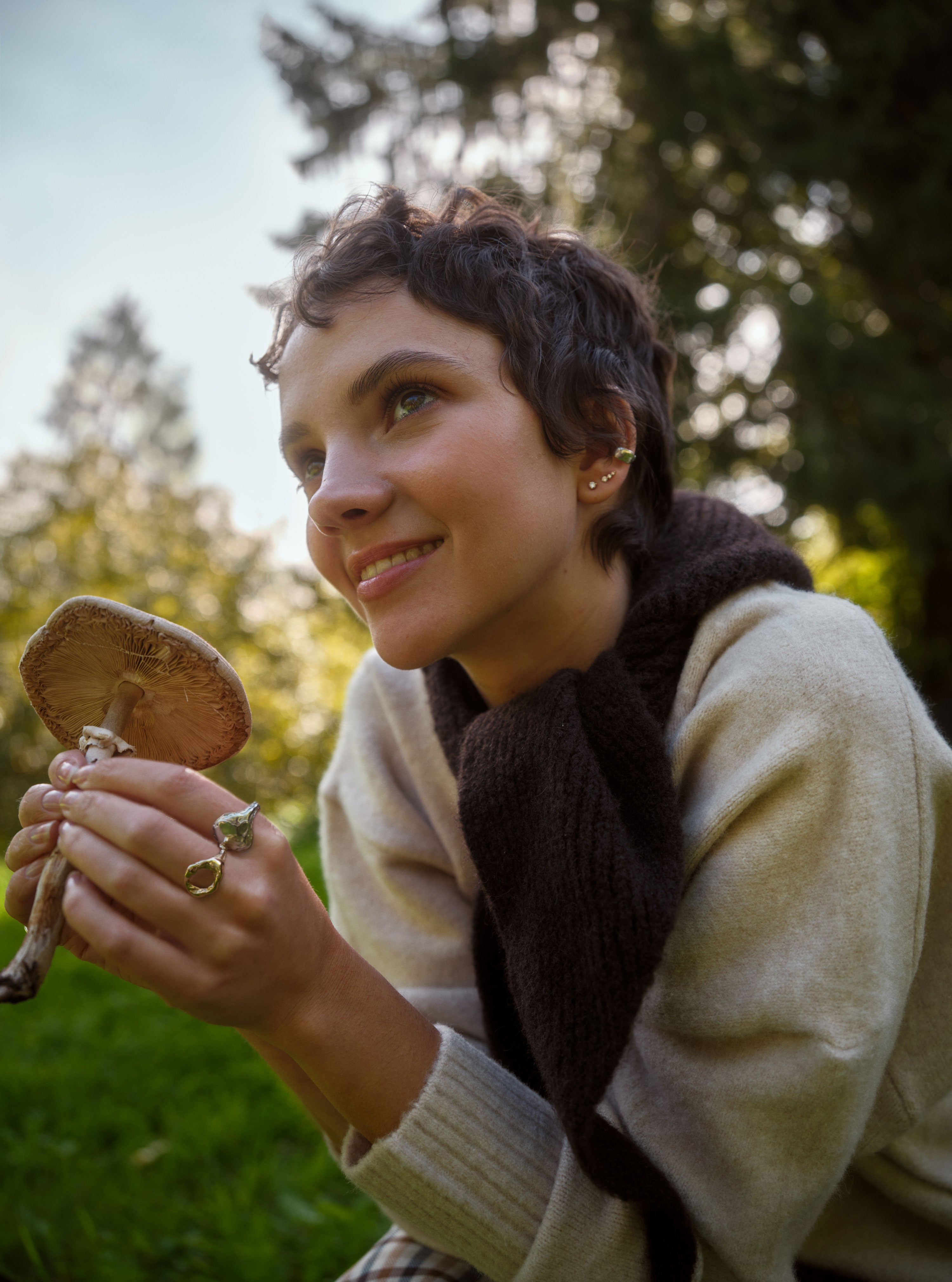 Woman wearing jewellery holding a mushroom outdoors with trees in the background