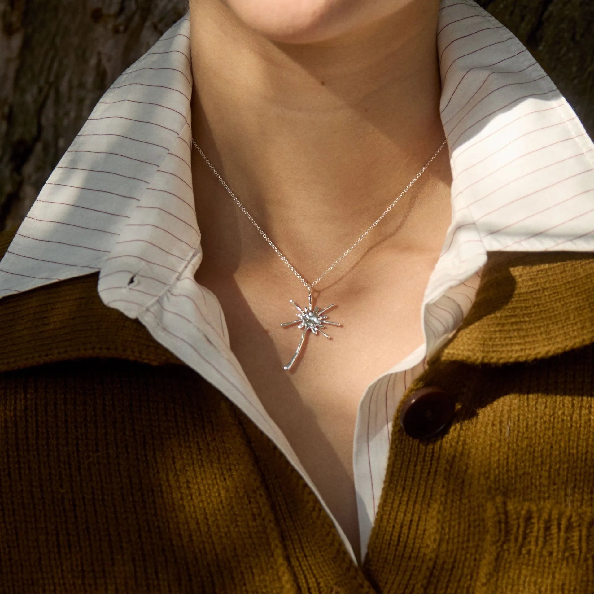 Person wearing a necklace with a dandelion-shaped pendant against a natural tree bark background
