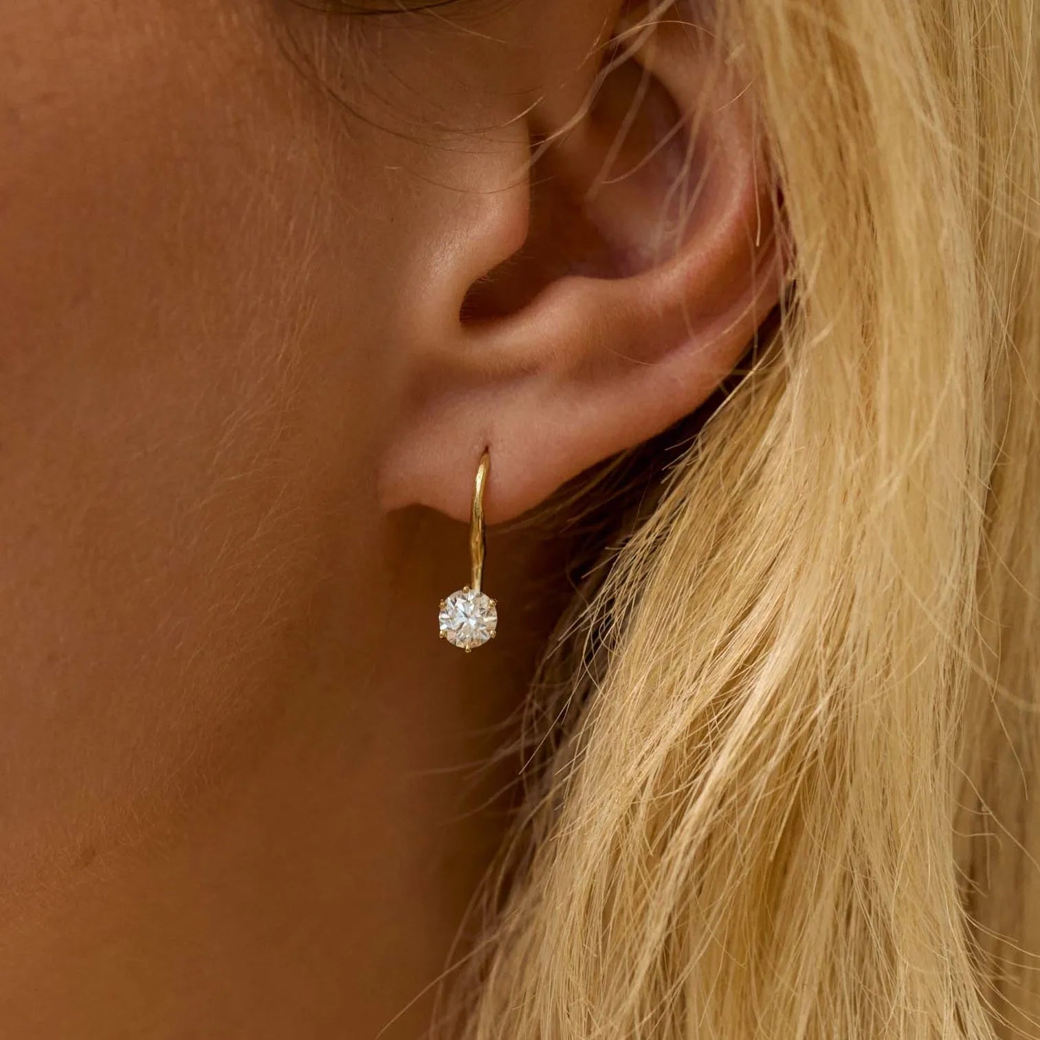 Close-up of an ear wearing a gold earring with a diamond, against a blurred background.