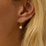 Close-up of an ear wearing a gold earring with a diamond, against a blurred background.