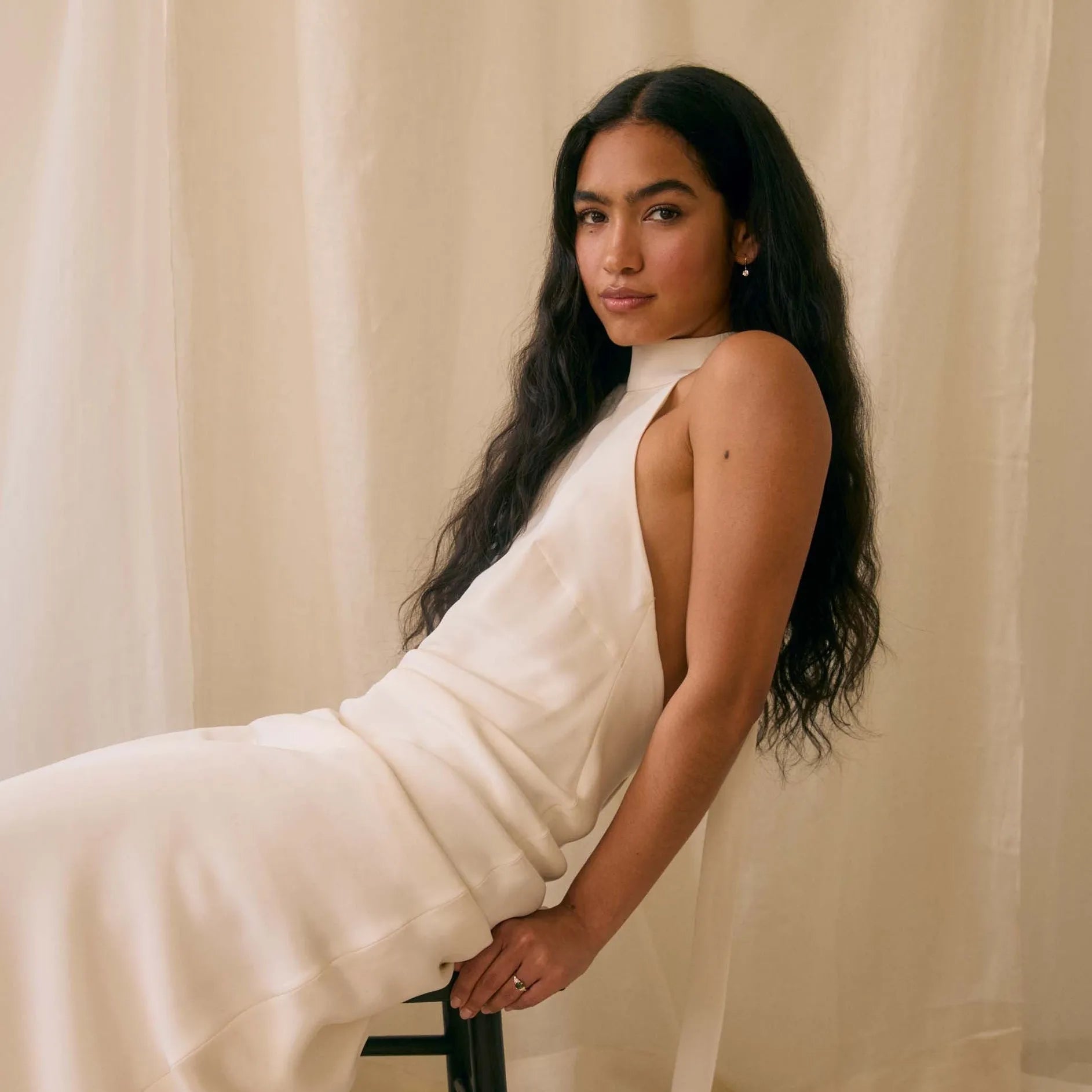 woman in white dress leaning back on a stool with a beige background, wearing diamond hook earrings and engagement ring