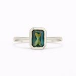 A white gold ring with a rectangular emerald-cut green gemstone featuring iridescent blue and gold reflections, set against a plain white background.