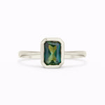 A white gold ring with a rectangular emerald-cut green gemstone featuring iridescent blue and gold reflections, set against a plain white background.