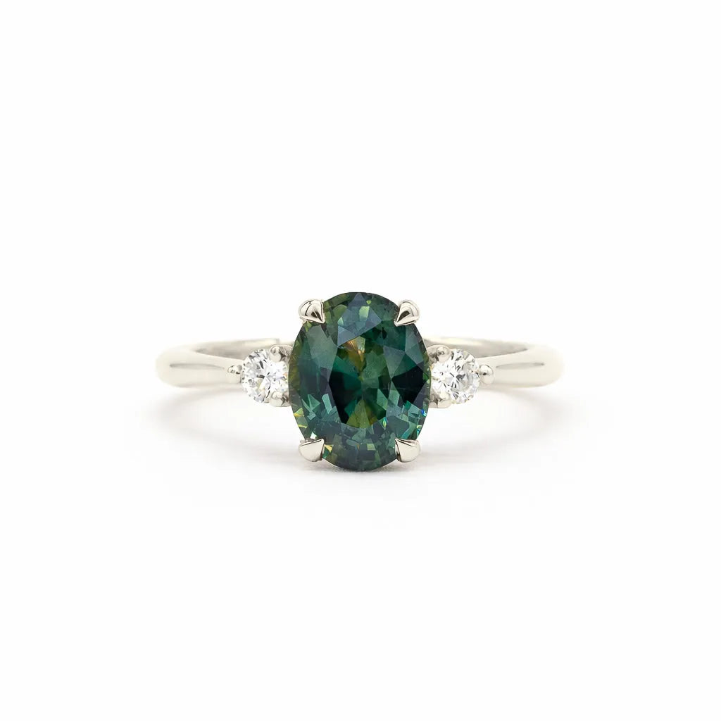 an oval-cut teal sapphire engagement ring with two round diamonds, set in white gold or platinum against a white background