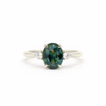 an oval-cut teal sapphire engagement ring with two round diamonds, set in white gold or platinum against a white background
