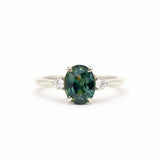 an oval-cut teal sapphire engagement ring with two round diamonds, set in white gold or platinum against a white background