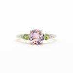 A white gold ring featuring a central cushion-cut light pink amethyst gemstone, flanked by two round green sapphires and small clear accent diamonds, set against a plain white background.