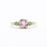 A white gold ring featuring a central cushion-cut light pink amethyst gemstone, flanked by two round green sapphires and small clear accent diamonds, set against a plain white background.