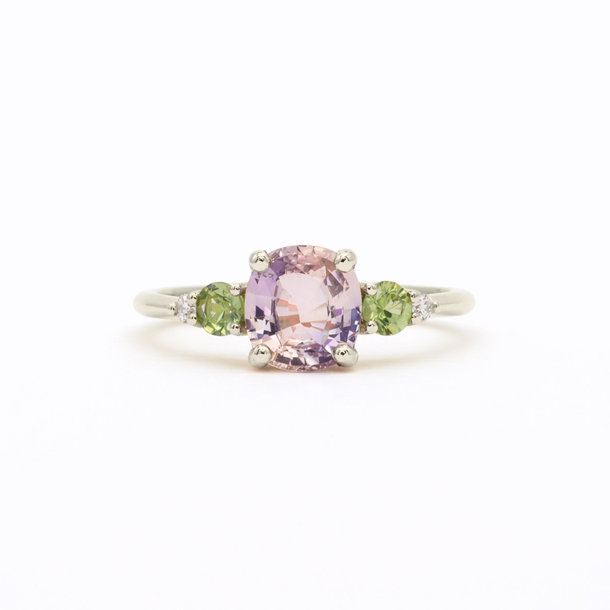 A white gold ring featuring a central cushion-cut light pink amethyst gemstone, flanked by two round green sapphires and small clear accent diamonds, set against a plain white background.