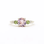 A white gold ring featuring a central cushion-cut light pink amethyst gemstone, flanked by two round green sapphires and small clear accent diamonds, set against a plain white background.