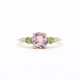 A white gold ring featuring a central cushion-cut light pink amethyst gemstone, flanked by two round green sapphires and small clear accent diamonds, set against a plain white background.