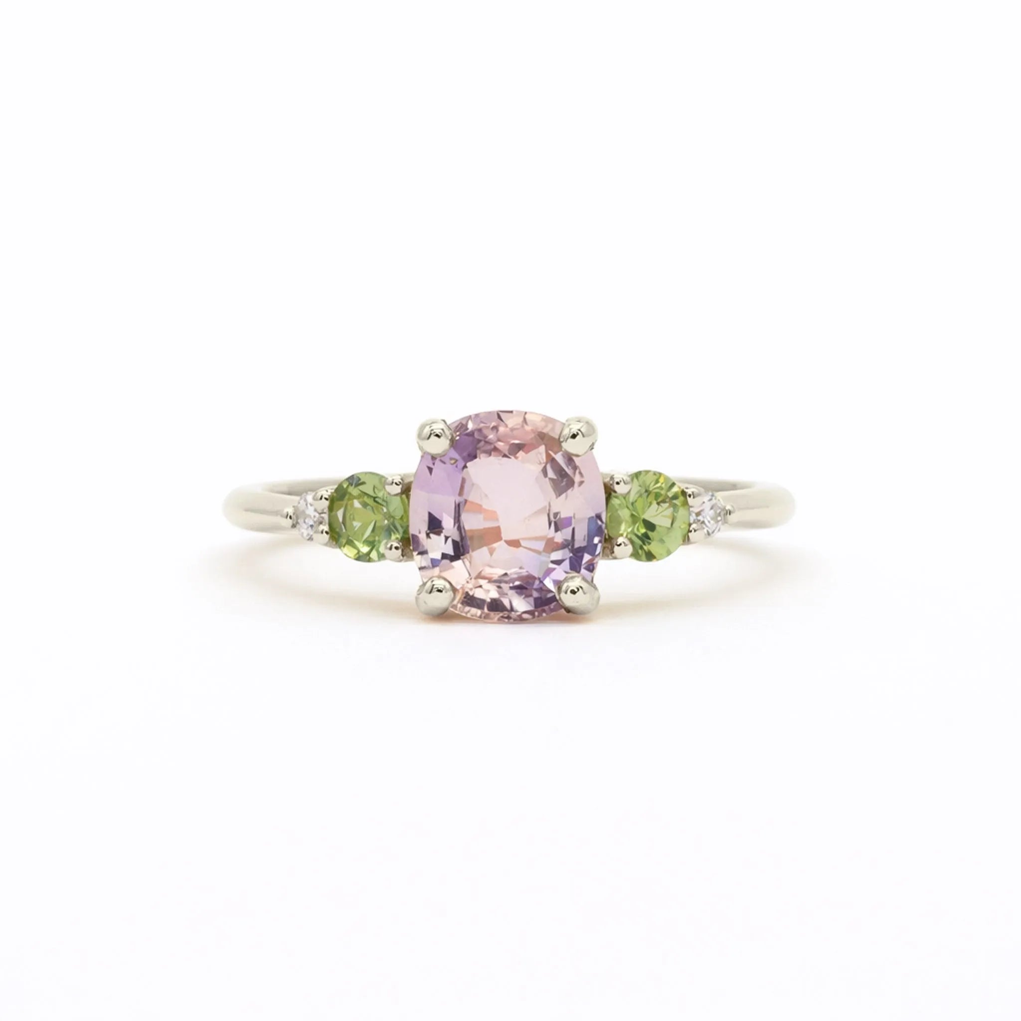 A white gold ring featuring a central cushion-cut light pink amethyst gemstone, flanked by two round green sapphires and small clear accent diamonds, set against a plain white background.