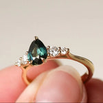 Gold ring with a greeny blue pear sapphire and round diamonds held in a hand against a neutral background