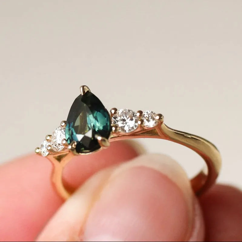 Gold ring with a greeny blue pear sapphire and round diamonds held in a hand against a neutral background