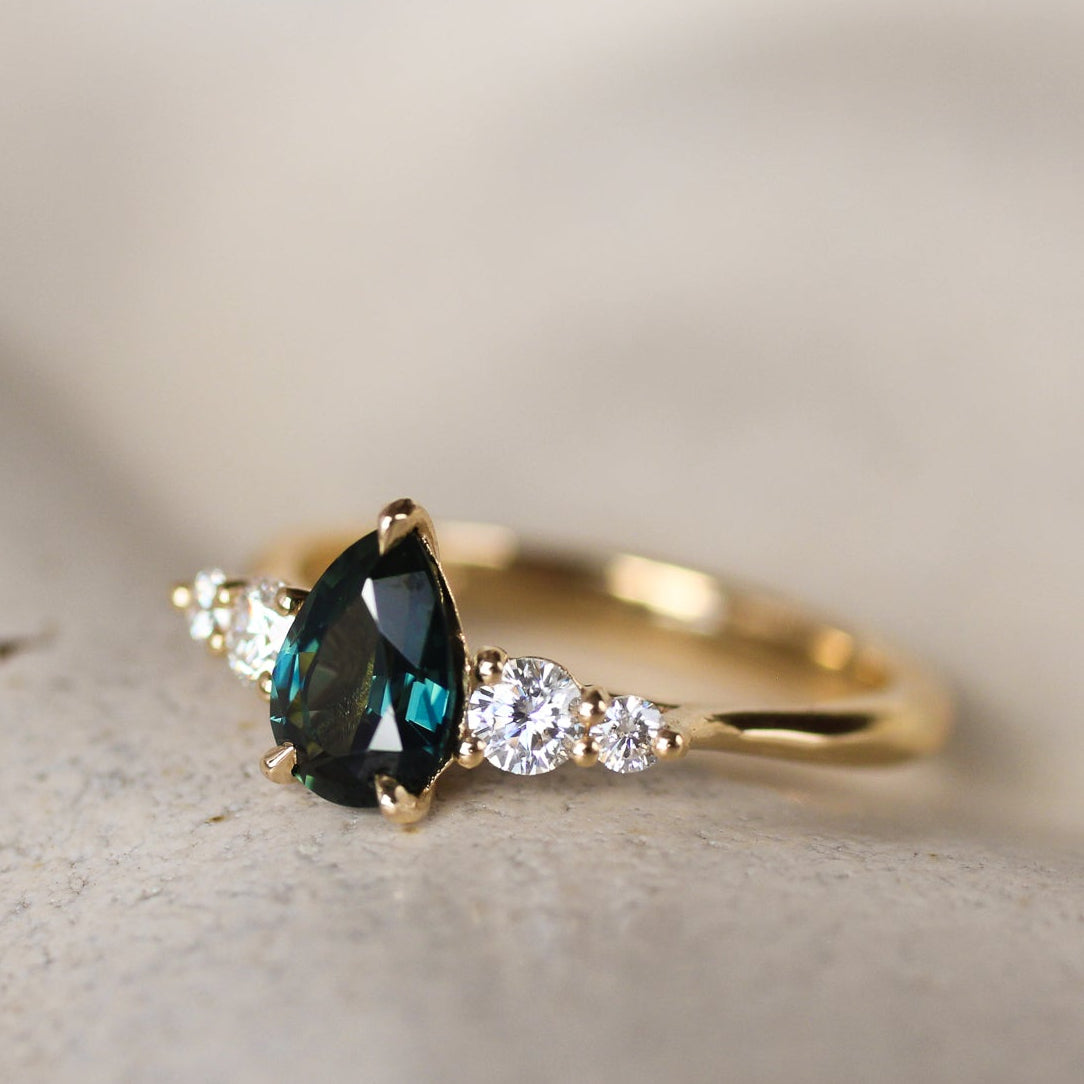 Gold ring with a deep green blue pear-shaped gemstone and smaller diamonds on a light background
