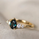 Gold ring with a deep green blue pear-shaped gemstone and smaller diamonds on a light background