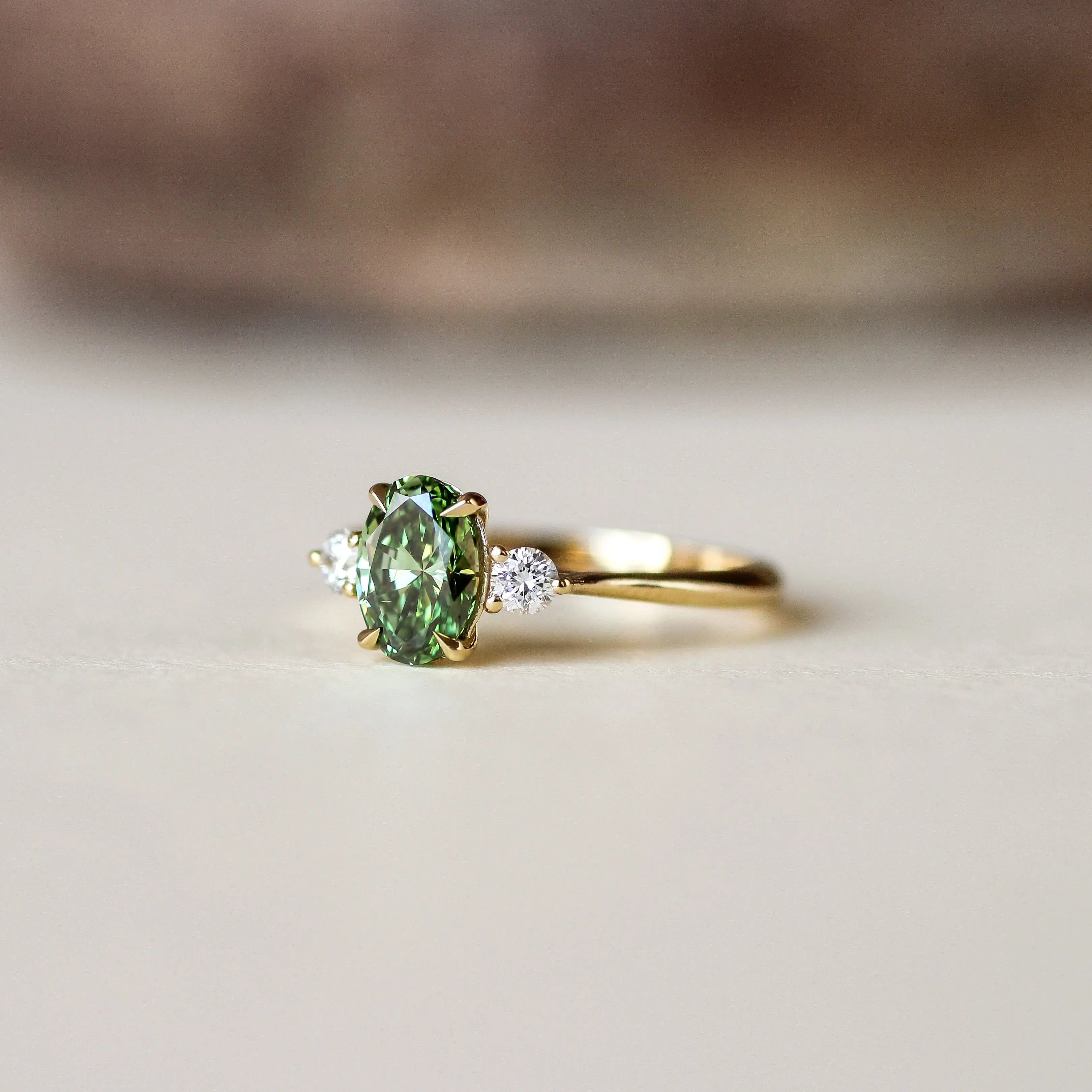 A gold ring featuring an oval-cut green gemstone center stone with two small clear accent stones on either side, resting on a light beige surface with a blurred brown background.