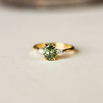 A gold ring featuring a central green gemstone and two small clear side stones, photographed on a light beige surface with a blurred brown background.