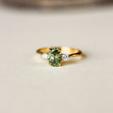 A gold ring featuring a central green gemstone and two small clear side stones, photographed on a light beige surface with a blurred brown background.