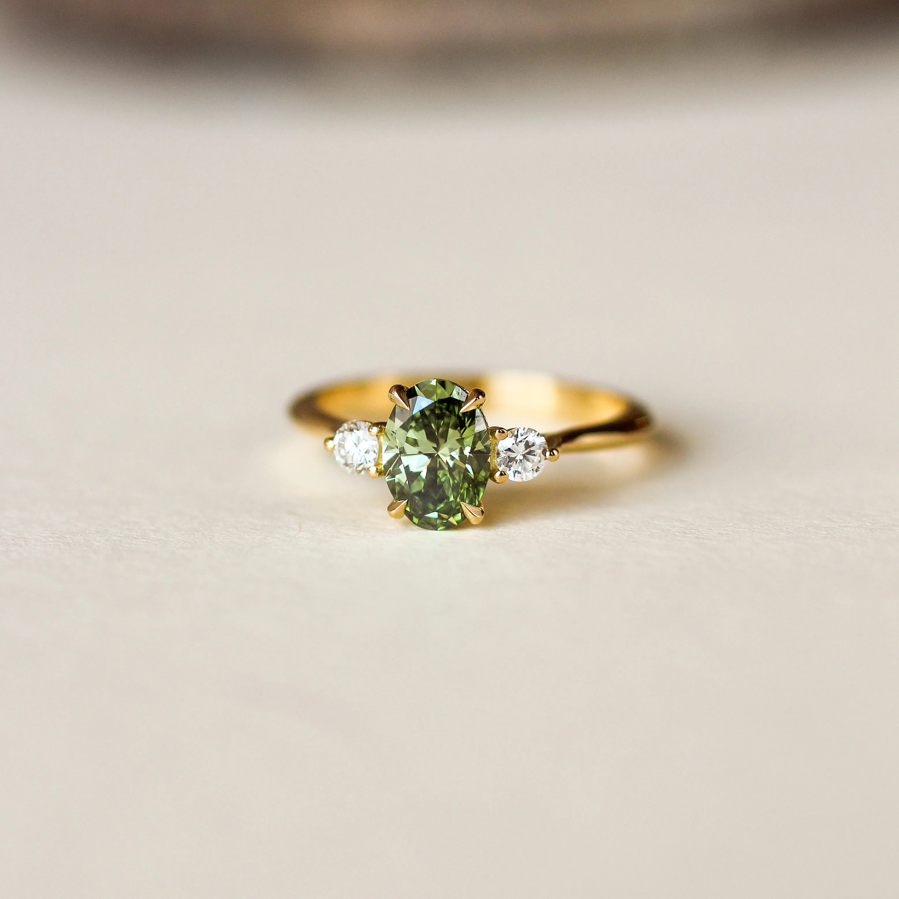 A gold ring featuring a central green gemstone and two small clear side stones, photographed on a light beige surface with a blurred brown background.