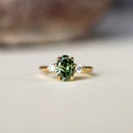 A gold ring featuring an oval-cut green diamond in the center, flanked by two small clear accent stones, resting on a light beige surface with a blurred background.