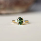 A gold ring featuring an oval-cut green diamond in the center, flanked by two small clear accent stones, resting on a light beige surface with a blurred background.
