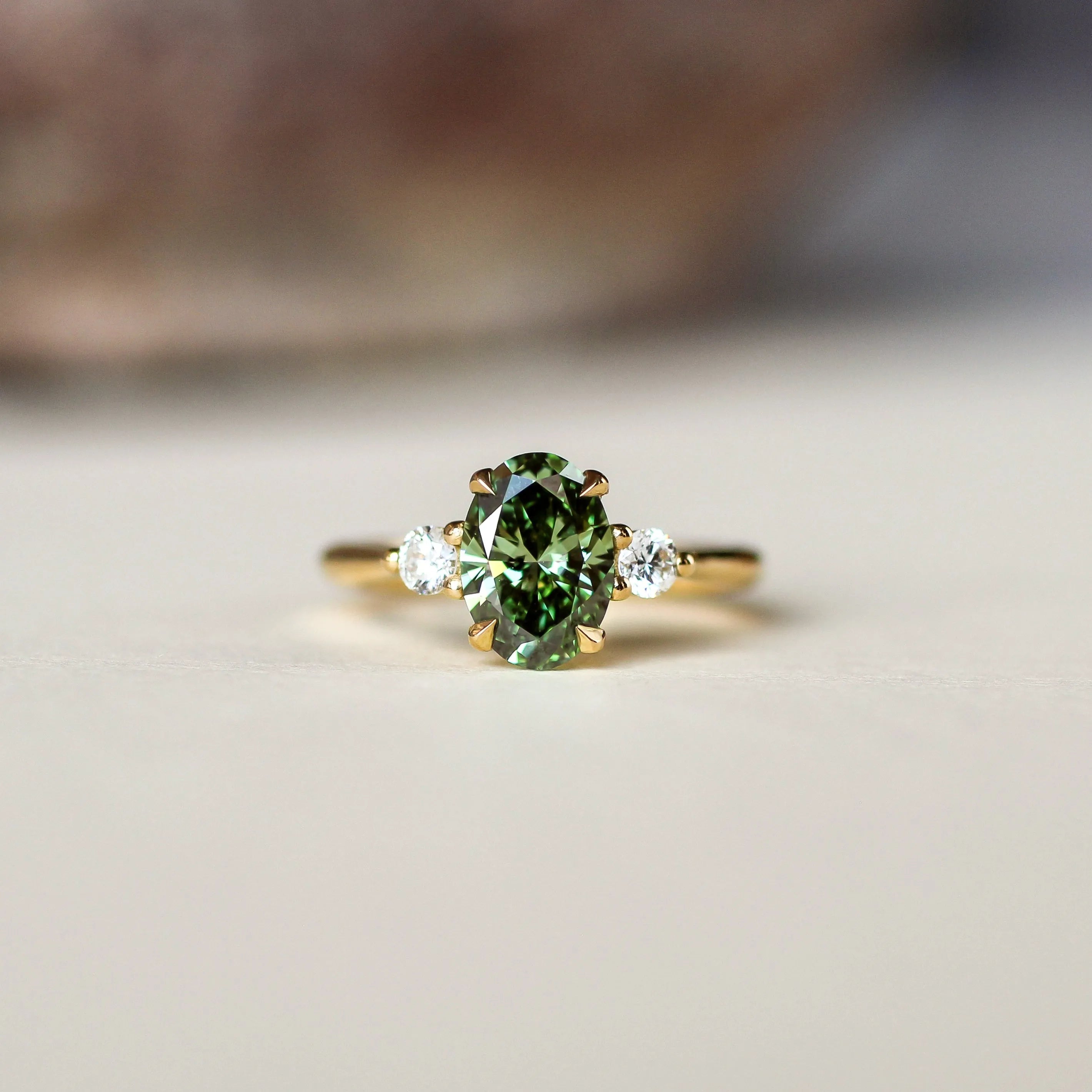 A gold ring featuring an oval-cut green diamond in the center, flanked by two small clear accent stones, resting on a light beige surface with a blurred background.