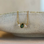A delicate gold necklace with a small pendant featuring a green oval sapphire and clear diamonds, resting on the rim of a textured ceramic dish.