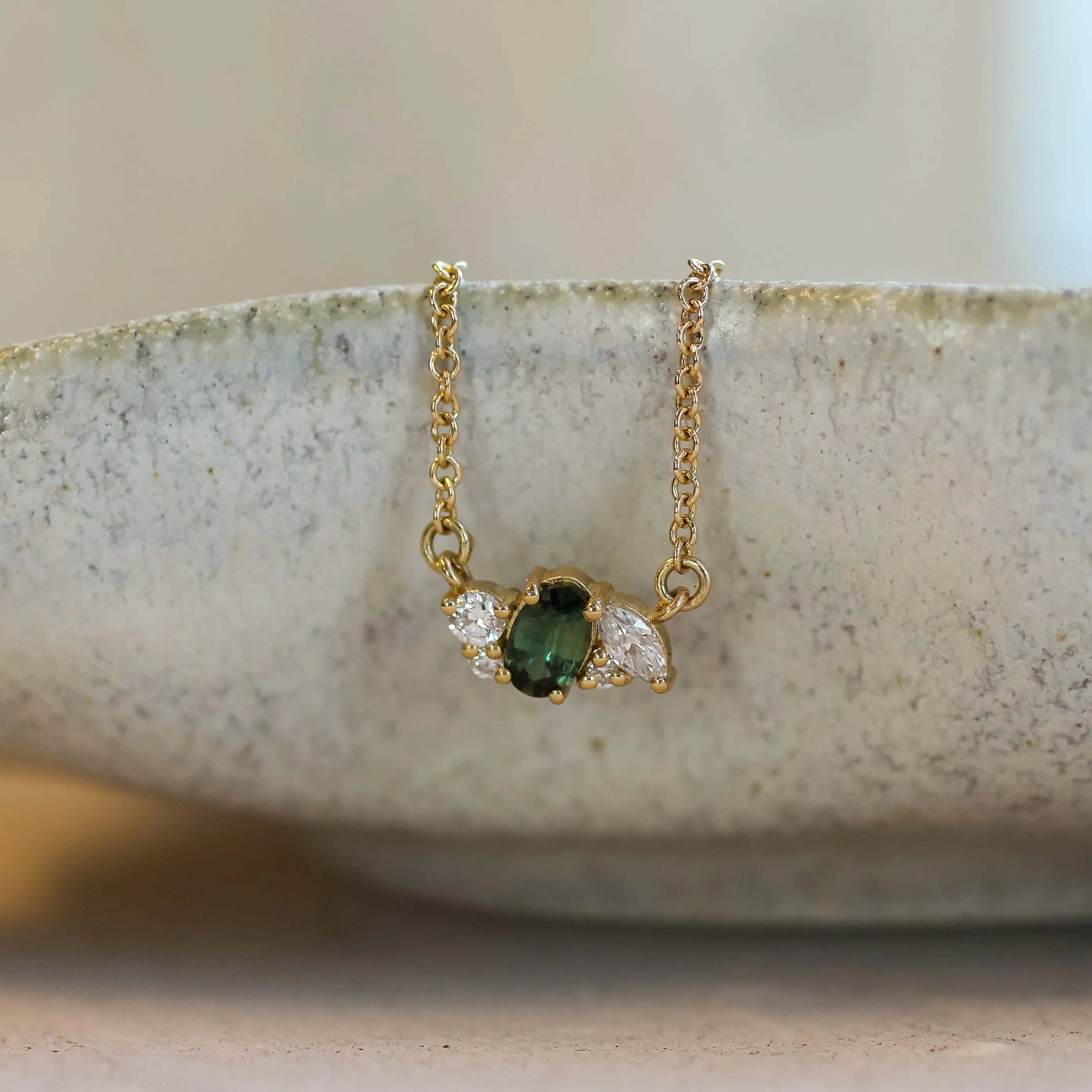 A delicate gold necklace with a small pendant featuring a green oval sapphire and clear diamonds, resting on the rim of a textured ceramic dish.