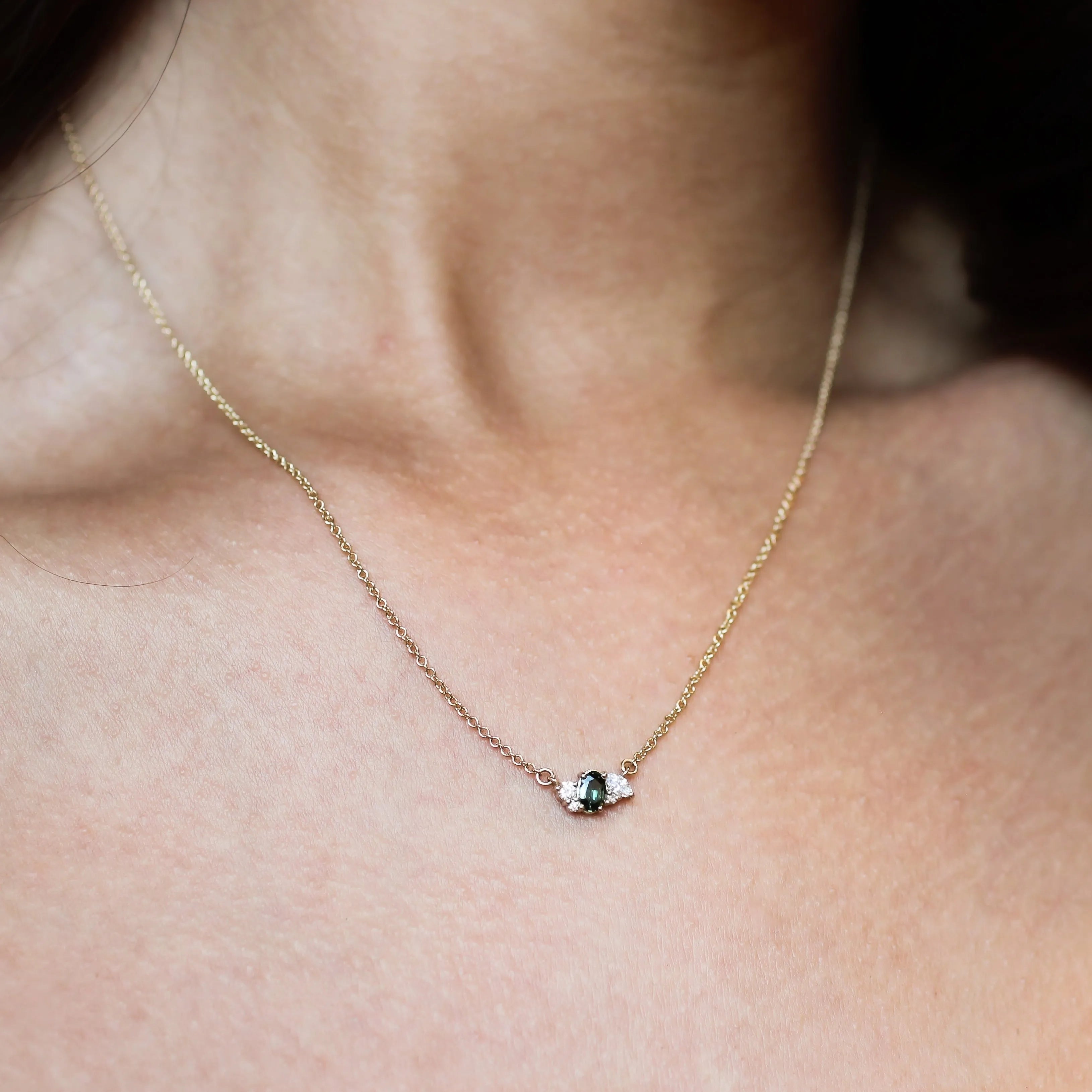 A delicate gold chain necklace with a small cluster of gemstones including a green sapphire and smaller diamonds, shown worn on a model's neck.