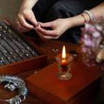 Person handling jewelry near a wooden box with rings and a lit candle on a table.